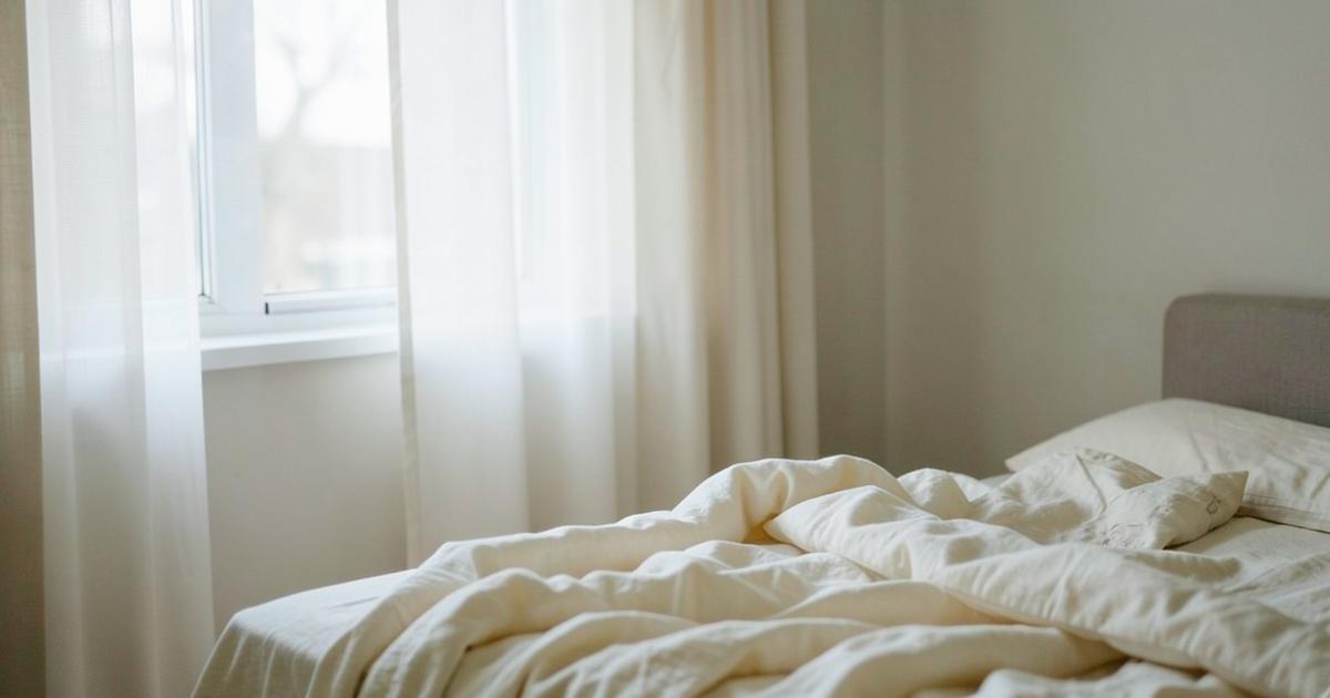 Soft morning light through sheer curtains falling across an unmade bed with cream linen