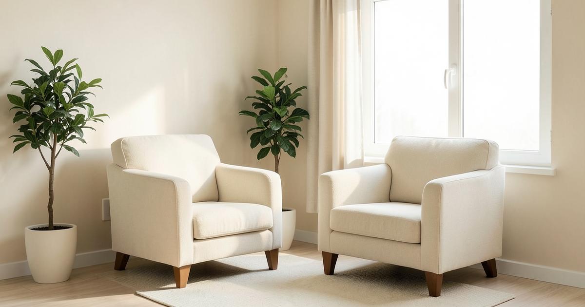Two empty cream armchairs beside a bright window in a calm, plant-filled counselling room